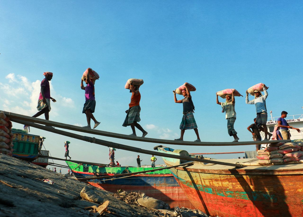 A group of laborers carrying sacks on a wooden plank at a busy dockside with colorful boats.
